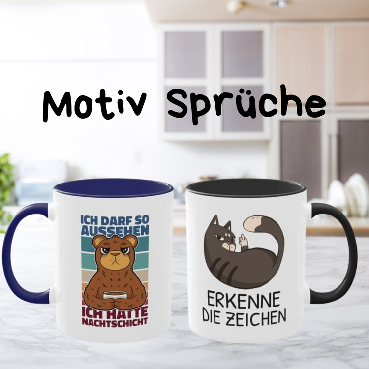 Zwei Motiv-Tassen: links eine mit einem mürrischen Bären und dem Spruch "Ich darf so aussehen, ich hatte Nachtschicht", rechts eine mit einer Katze und dem Spruch "Erkenne die Zeichen". Hintergrund: Küche